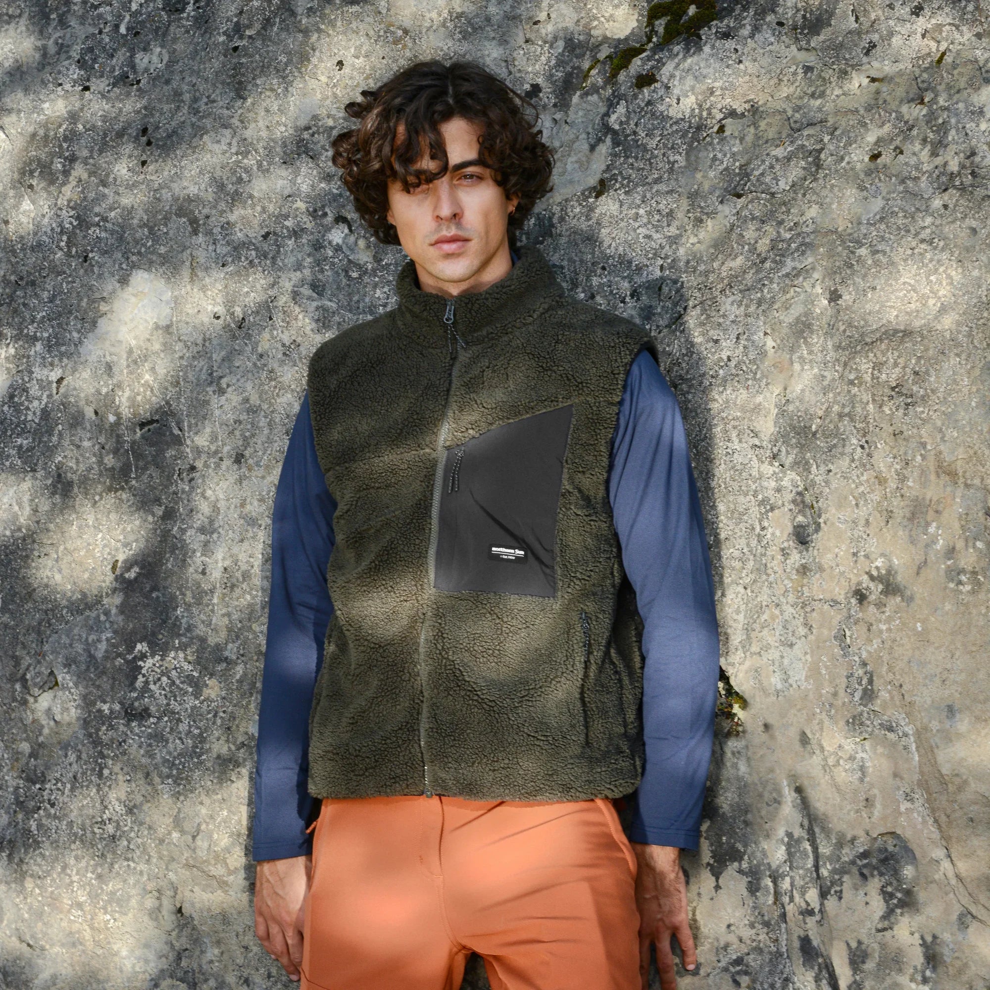 Man wearing olive green sherpa vest, blue shirt, and orange pants against a stone wall