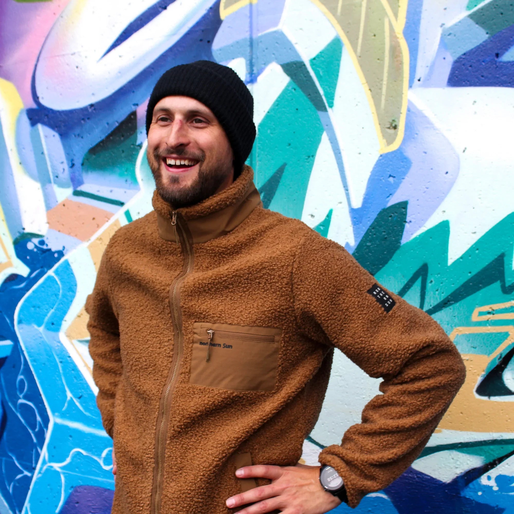Man wearing brown teddy fleece jacket and black beanie in front of colorful graffiti wall