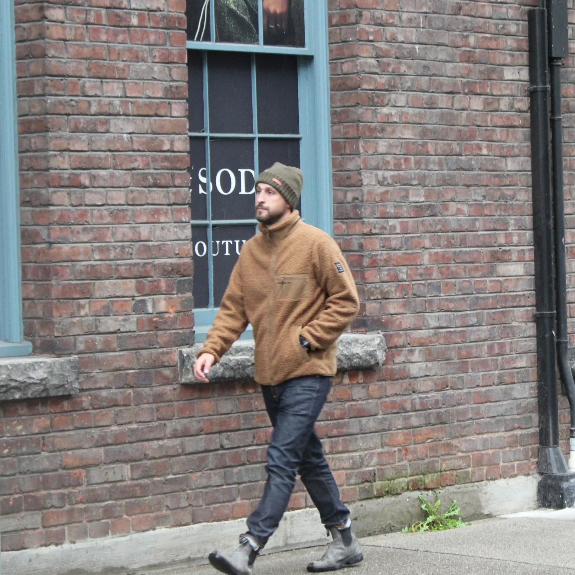 Man wearing brown teddy fleece jacket and beanie walking by brick building outdoors