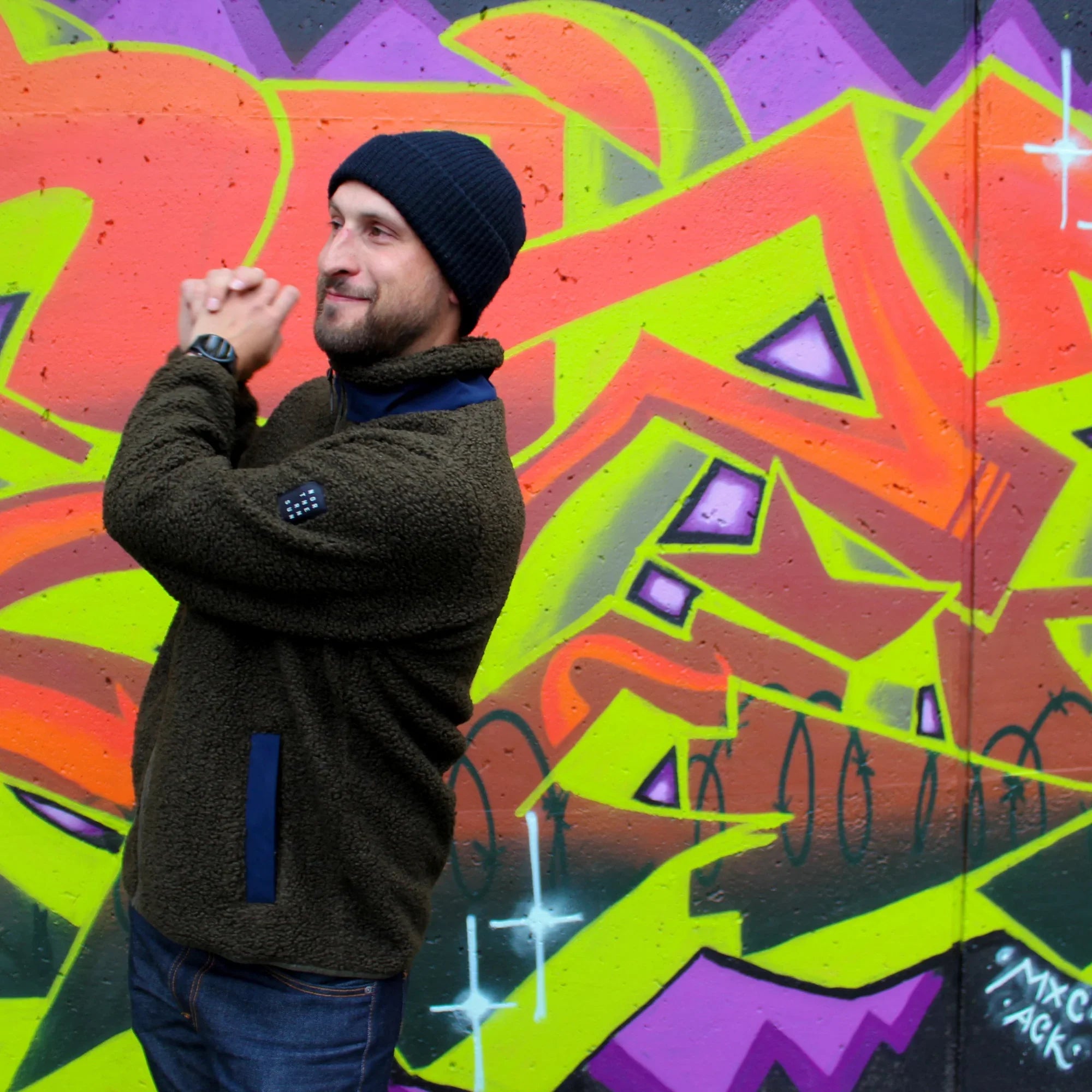 Man in green teddy fleece and black beanie posing by colorful graffiti wall