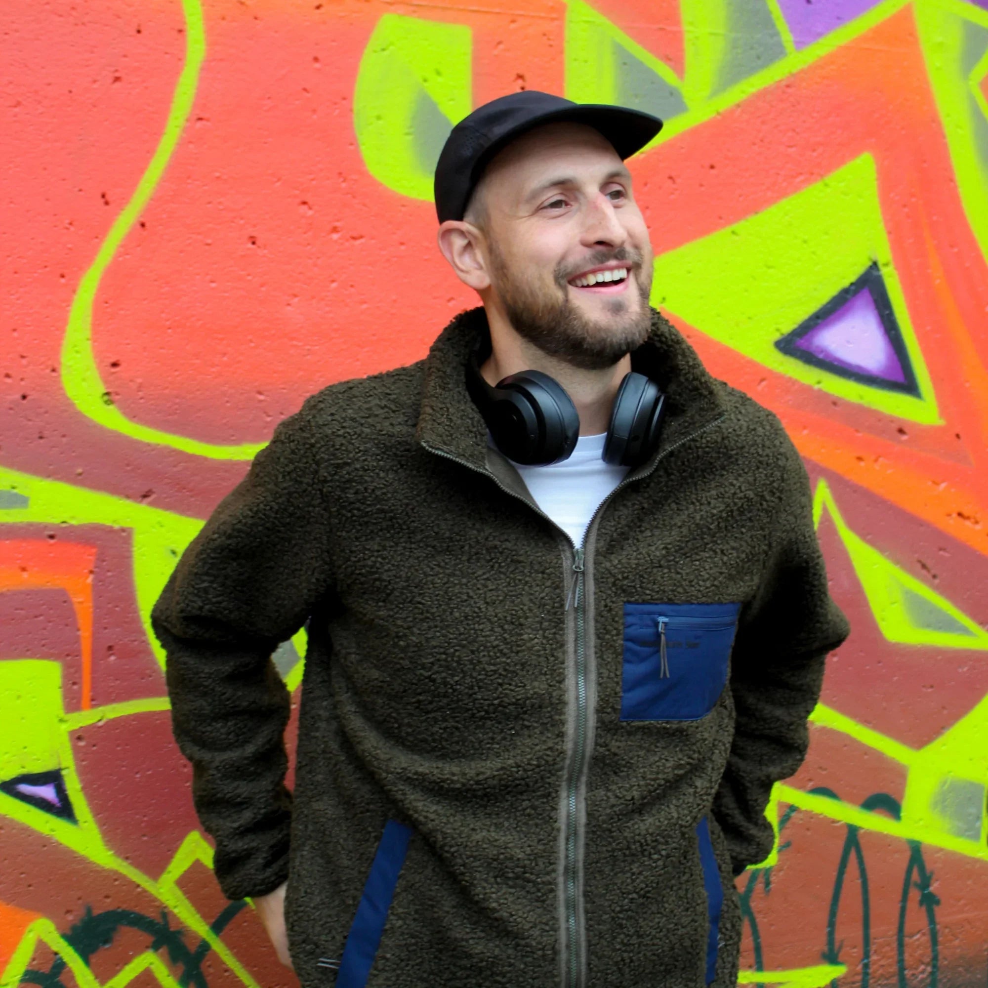 Man in green teddy fleece jacket with headphones in front of colorful city graffiti wall