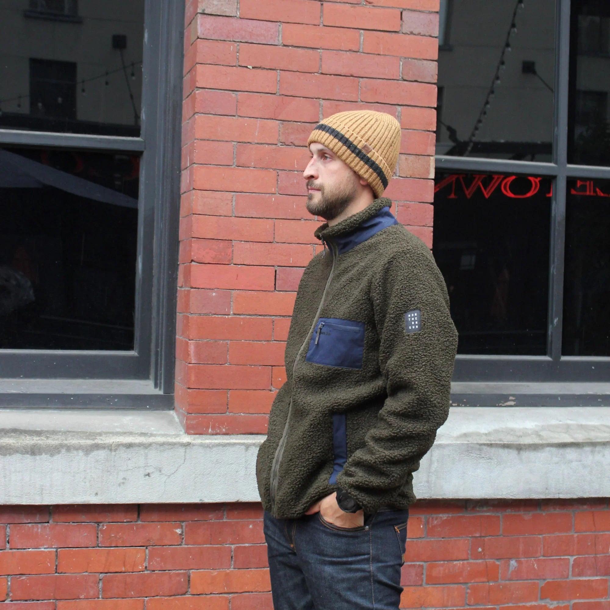Man wearing olive teddy fleece jacket and beanie standing by brick wall outdoors