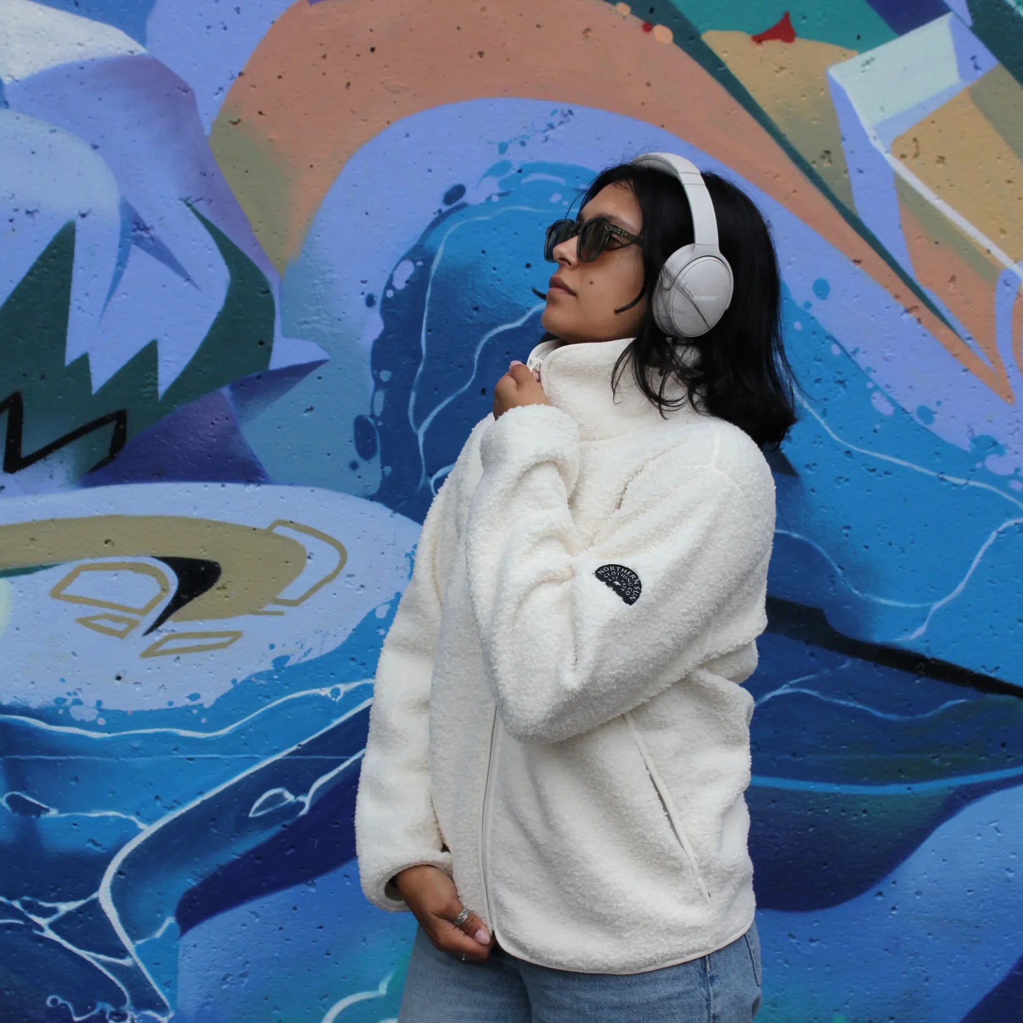Woman in white teddy fleece jacket and headphones standing by blue graffiti wall
