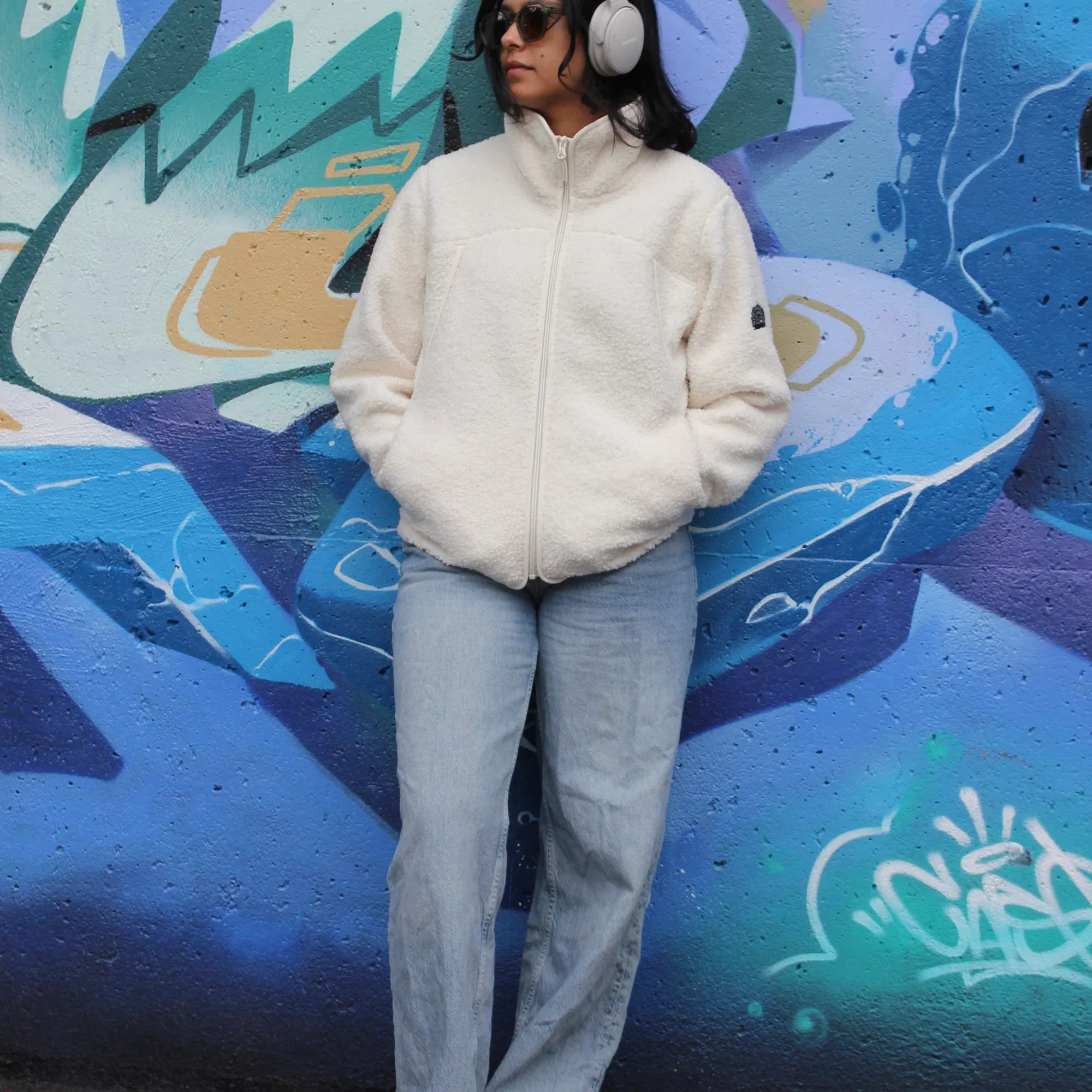 Woman in white teddy fleece jacket and jeans posing by blue graffiti wall