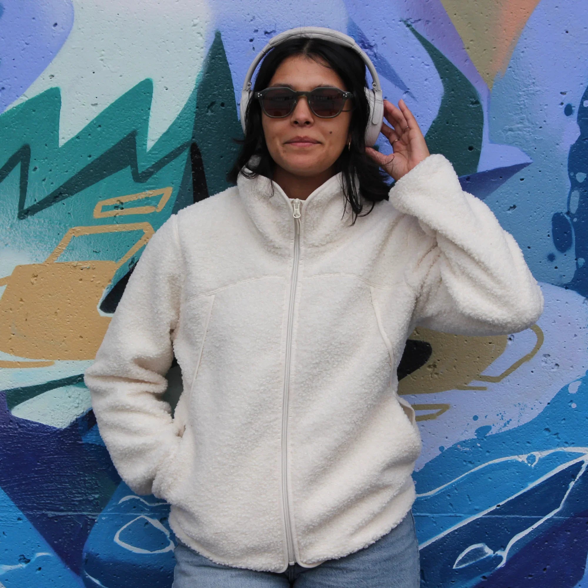Woman in white teddy fleece jacket with pockets, sunglasses, headphones, colorful graffiti wall