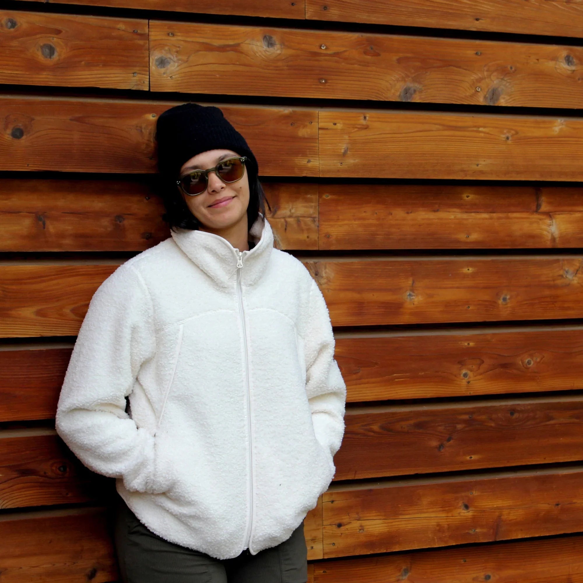 Woman in white teddy fleece jacket and beanie, standing by wooden wall outdoors