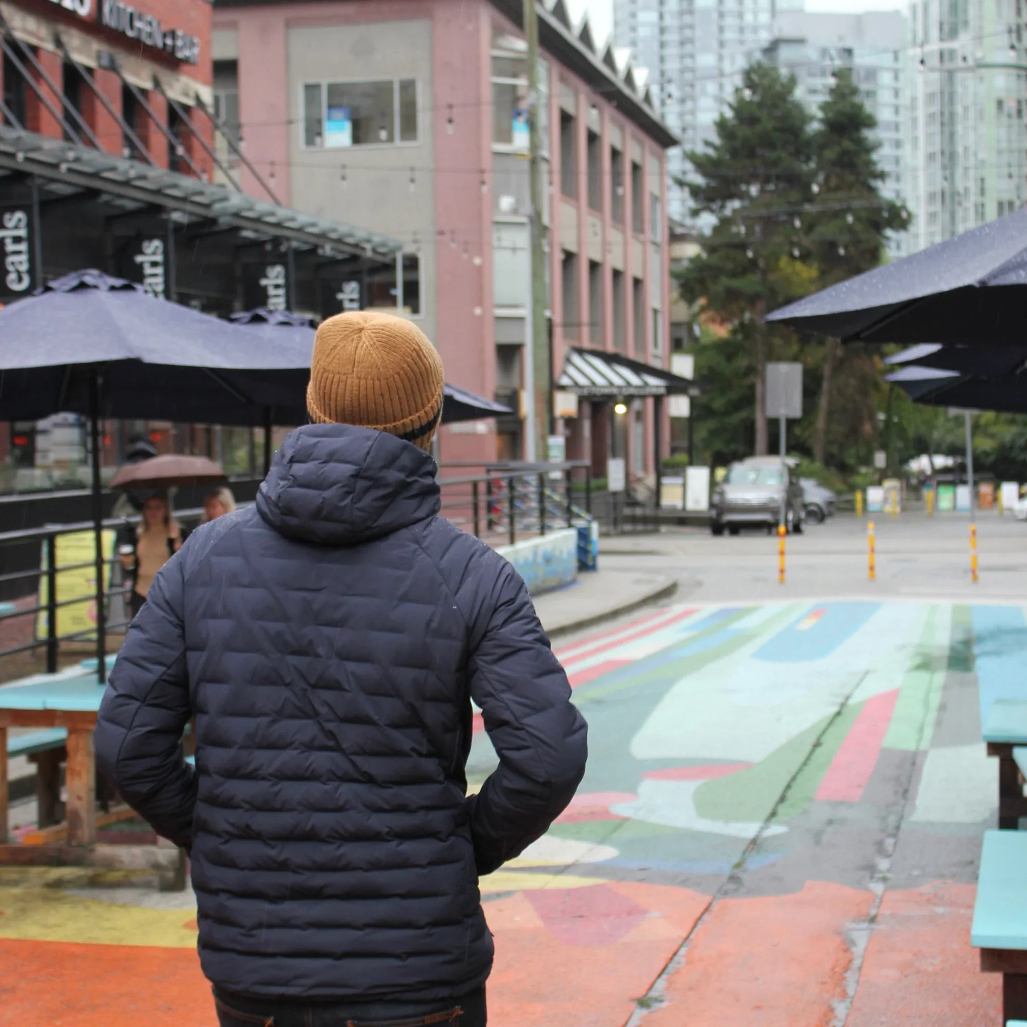 Man in navy Dawson down jacket and brown beanie on colorful urban street, city buildings