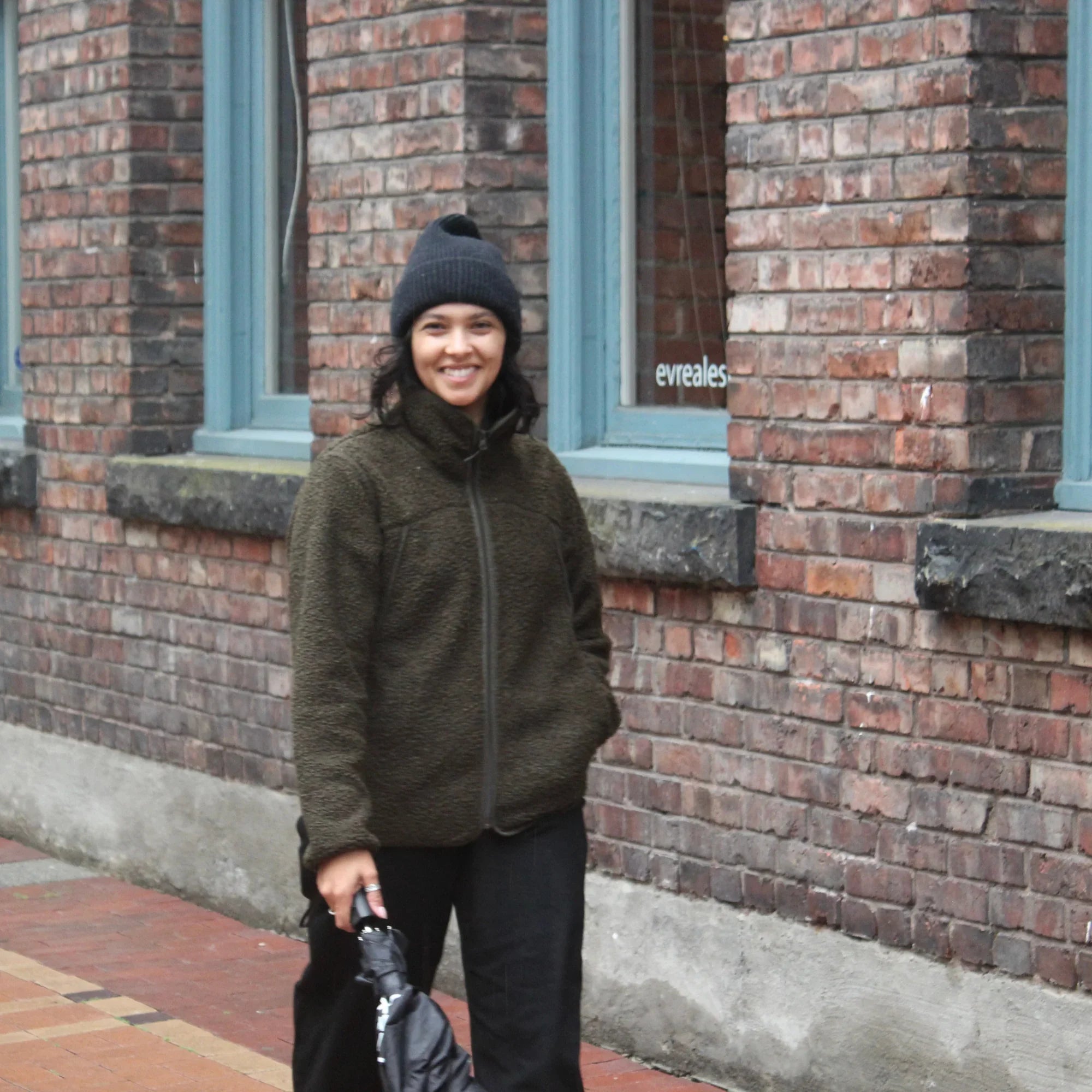 Smiling woman in dark teddy fleece jacket and beanie standing by urban brick wall
