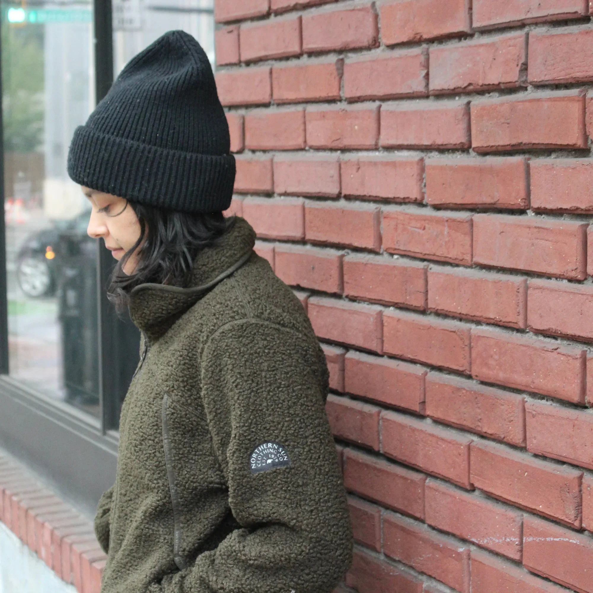 Woman in olive teddy fleece jacket and black beanie leaning on red brick wall city street