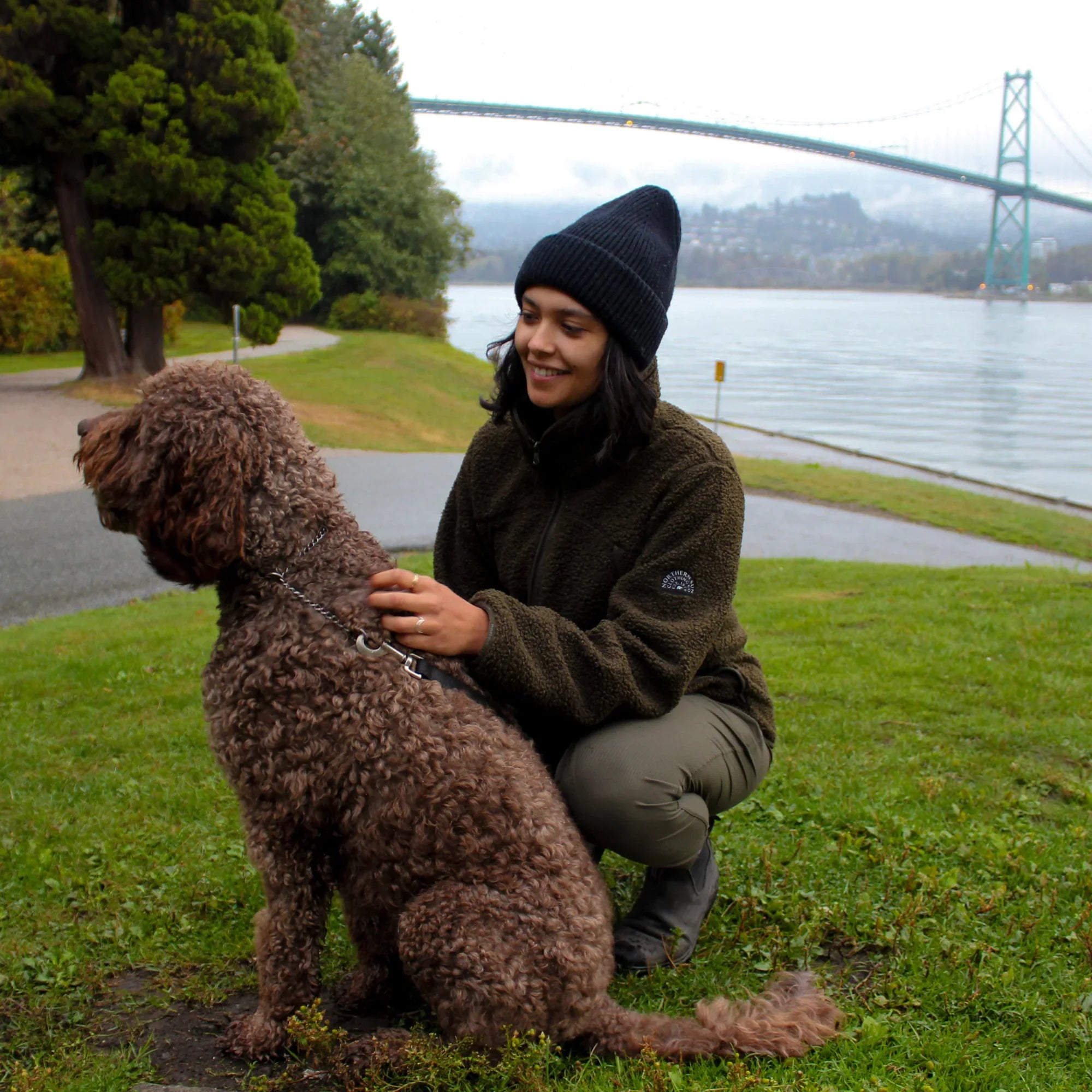 Woman in teddy fleece jacket walking brown curly dog near water and bridge in park