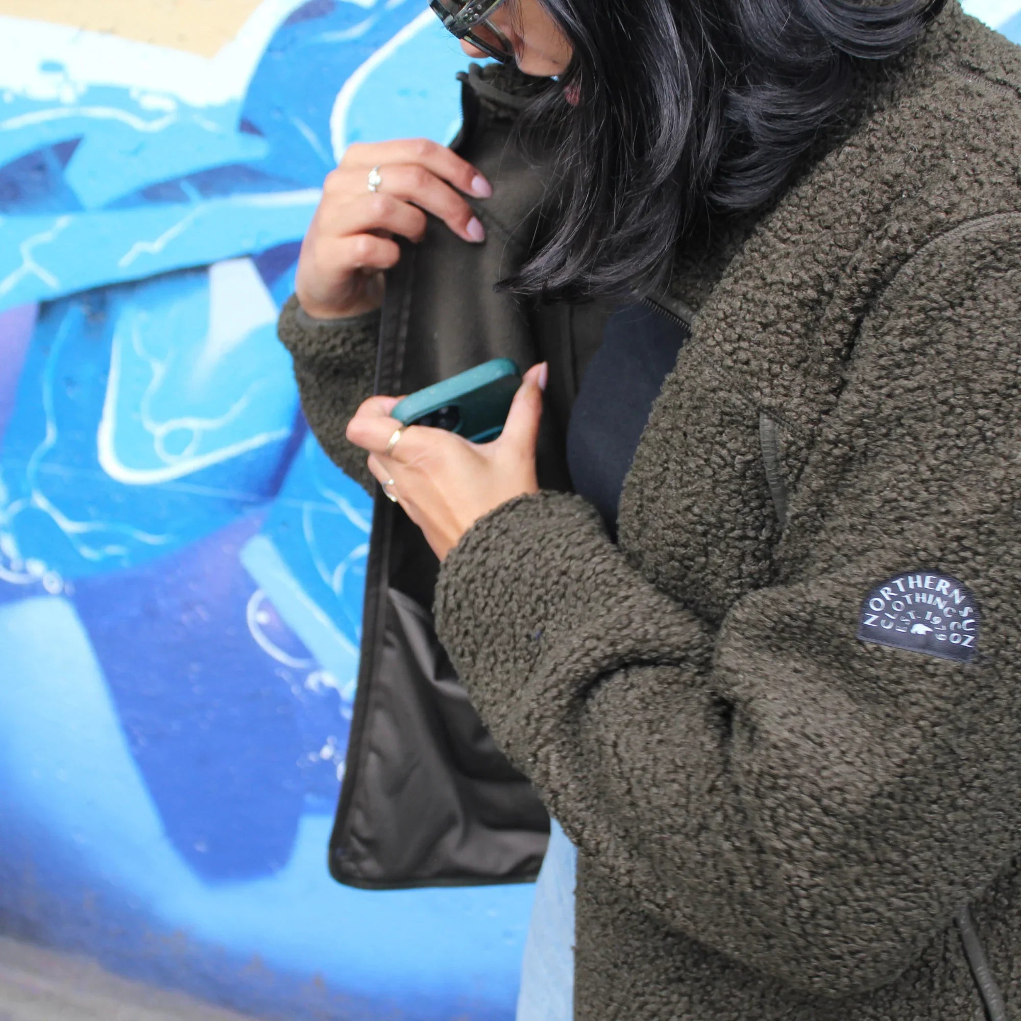 Woman wearing olive teddy fleece jacket with inside pockets, Northern Sun patch, blue mural background
