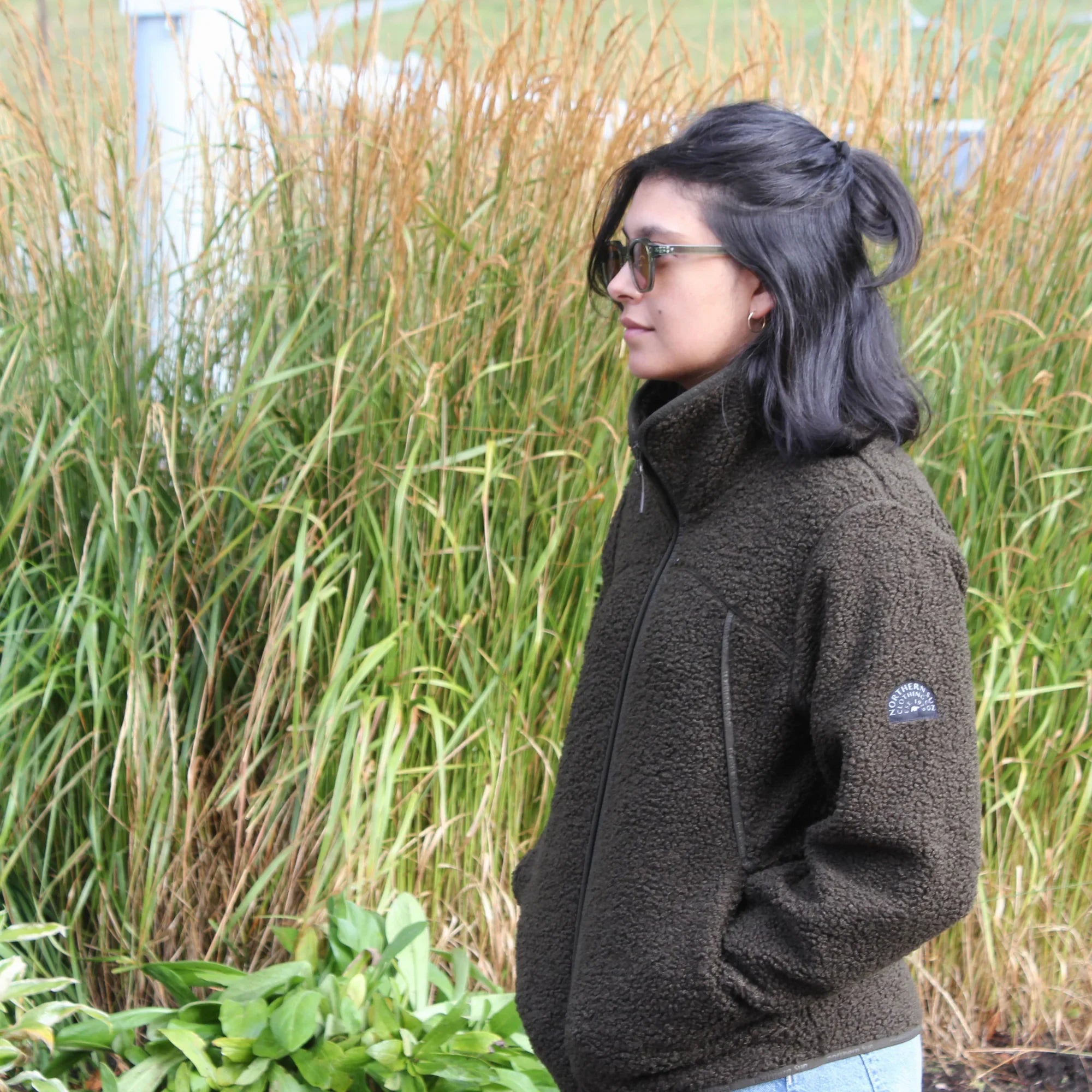 Woman wearing dark green teddy fleece jacket outdoors near tall grass, sunglasses on