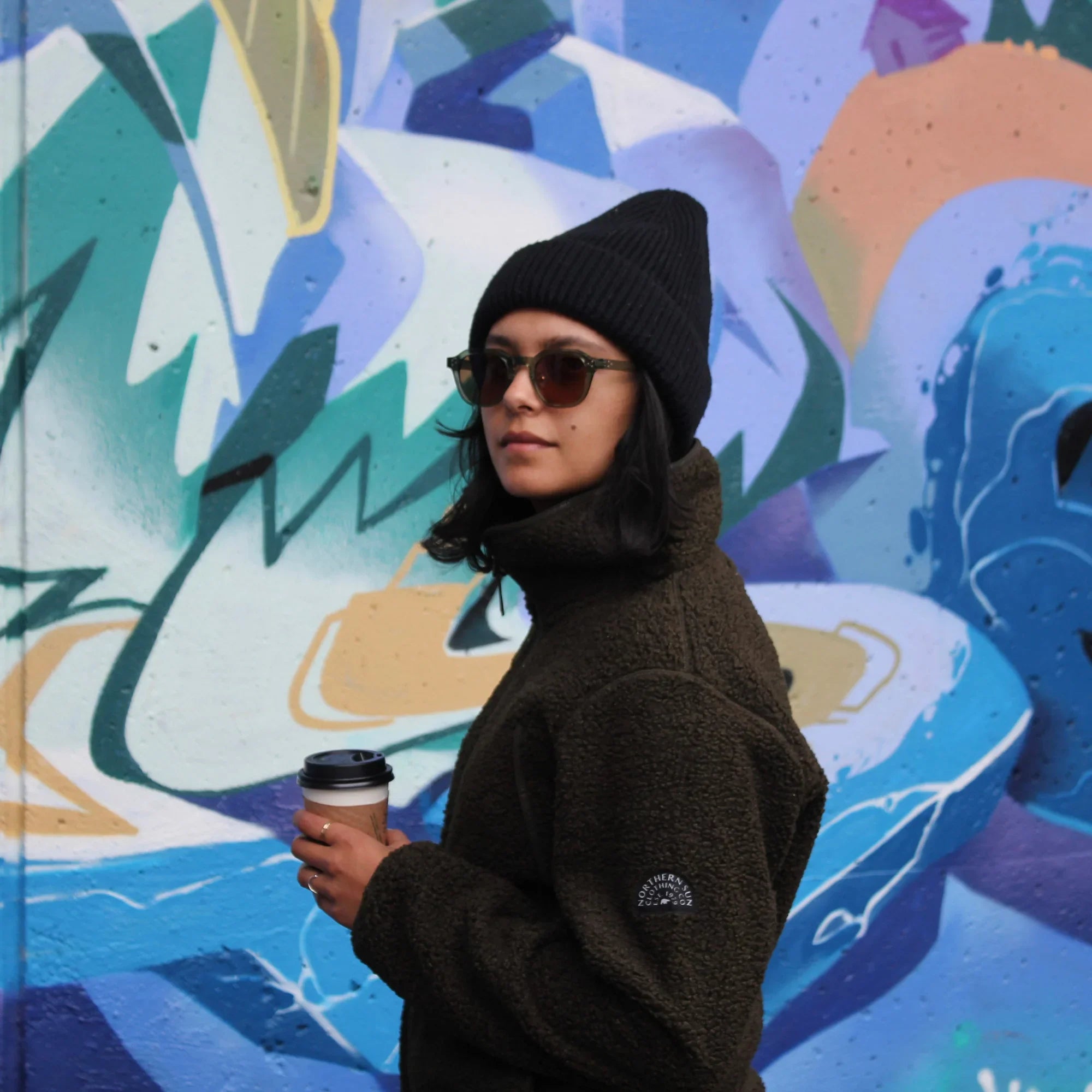 Woman in brown teddy fleece, sunglasses and beanie holding coffee, graffiti background