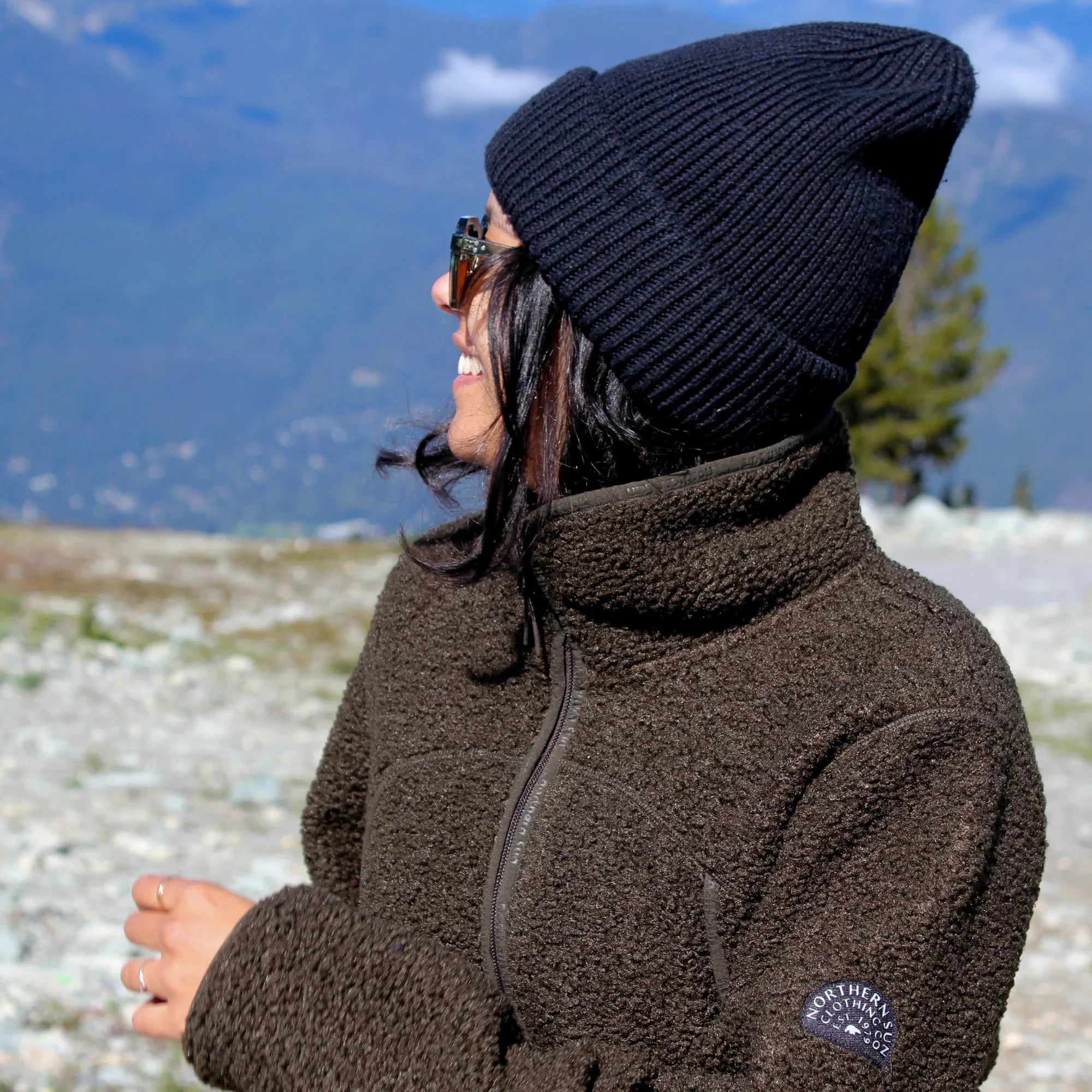 Woman wearing brown teddy fleece jacket and black knit beanie in green mountain landscape