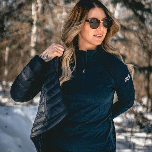 Woman wearing NorthWool quarter zip jacket, outdoors in snowy forest, putting on coat