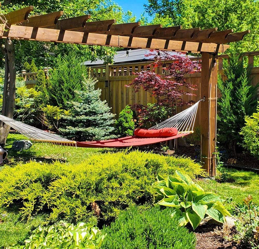 Red hammock under wooden pergola in lush green backyard garden with trees and shrubs