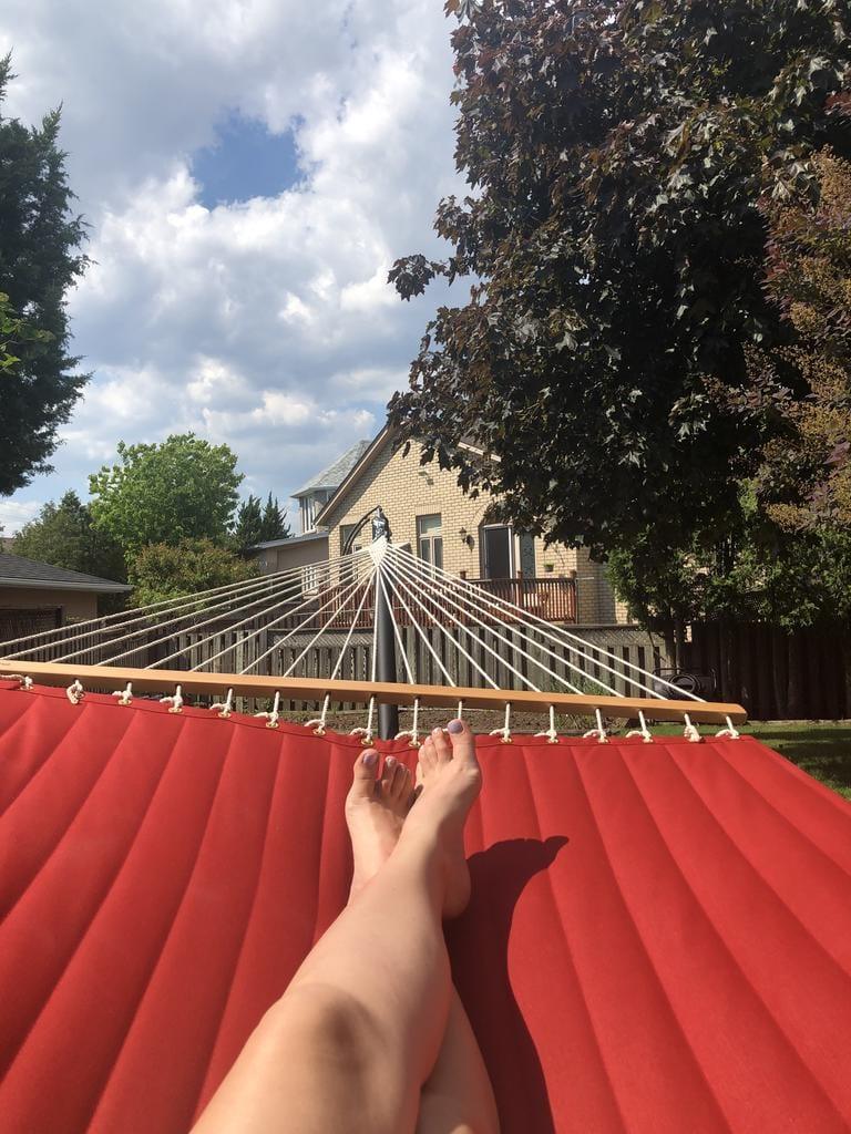 Person relaxing on red hammock in backyard with house and trees under blue sky