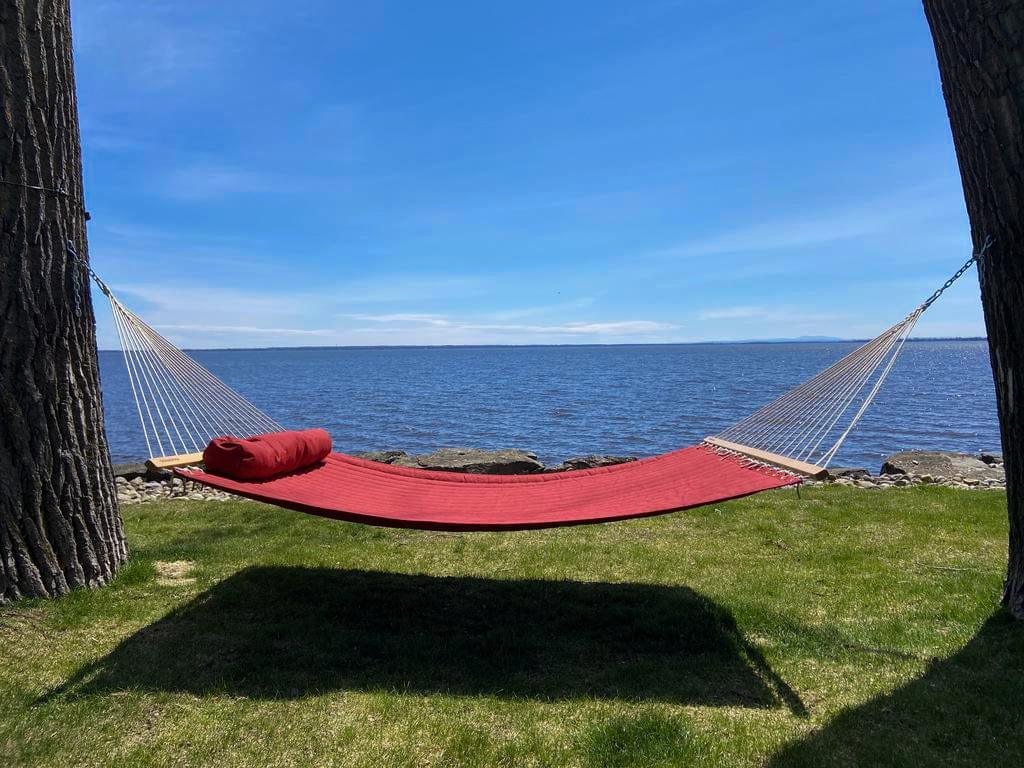 Red double quilted hammock with pillow hanging between trees by lake under blue sky