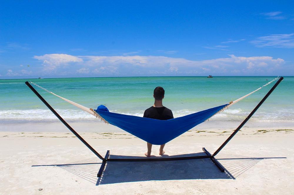 Man relaxing on a blue quilted hammock with stand on a sandy beach by the ocean