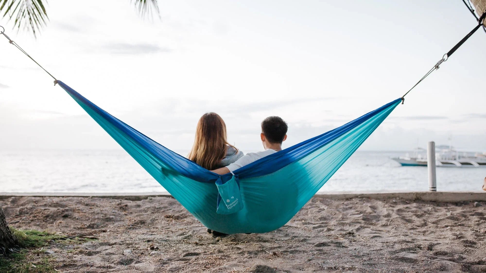 Couple relaxing in blue hammock on sandy beach with ocean and boats in background