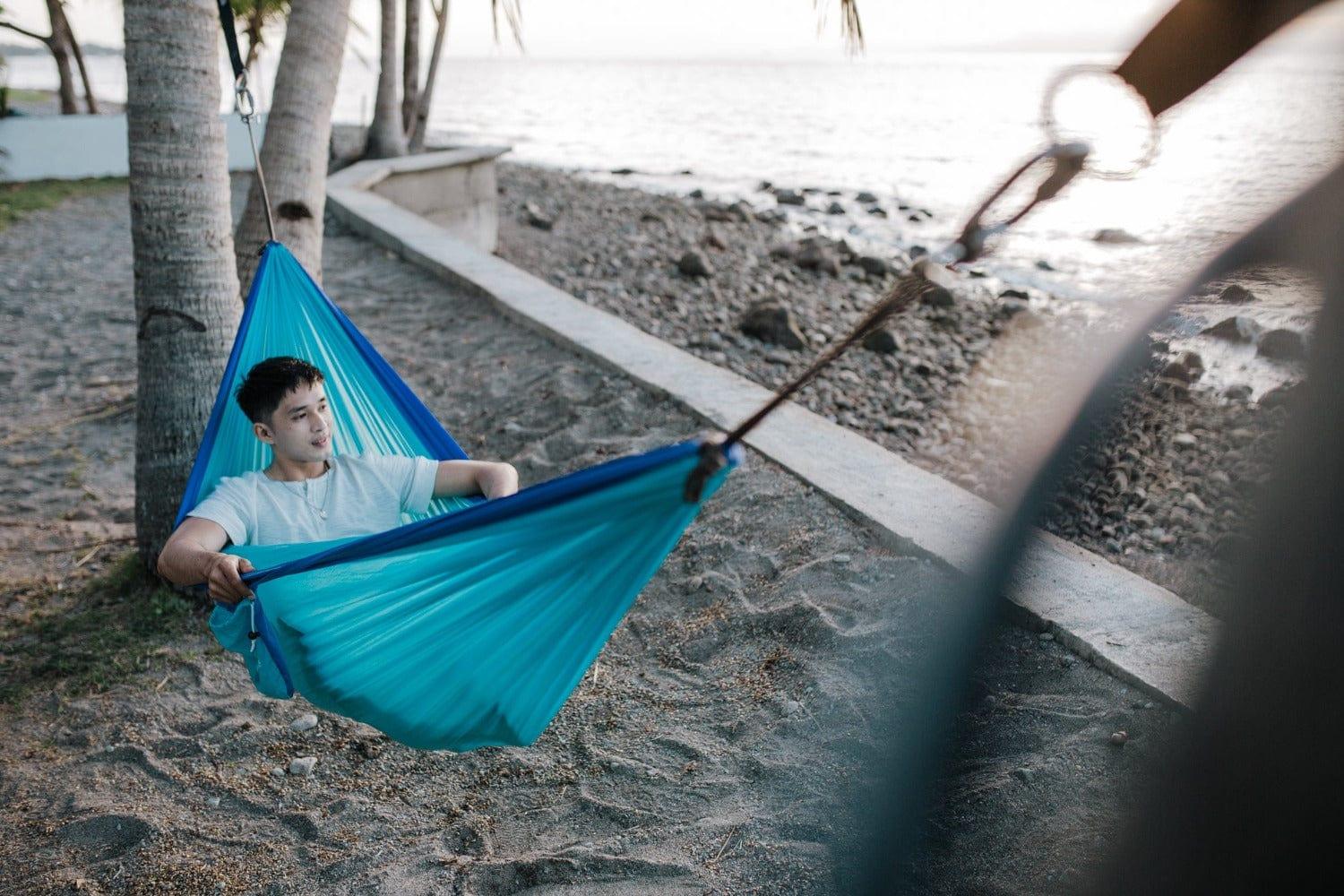 Man relaxing in a blue hammock between palm trees on a sandy beach at sunset
