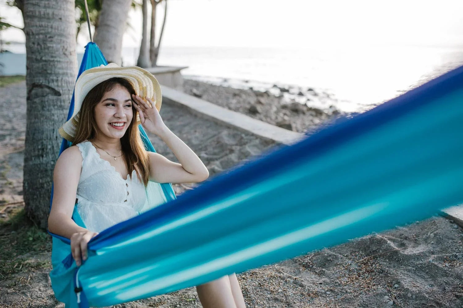 Smiling woman in a white dress and sunhat relaxing in a blue hammock by the beach