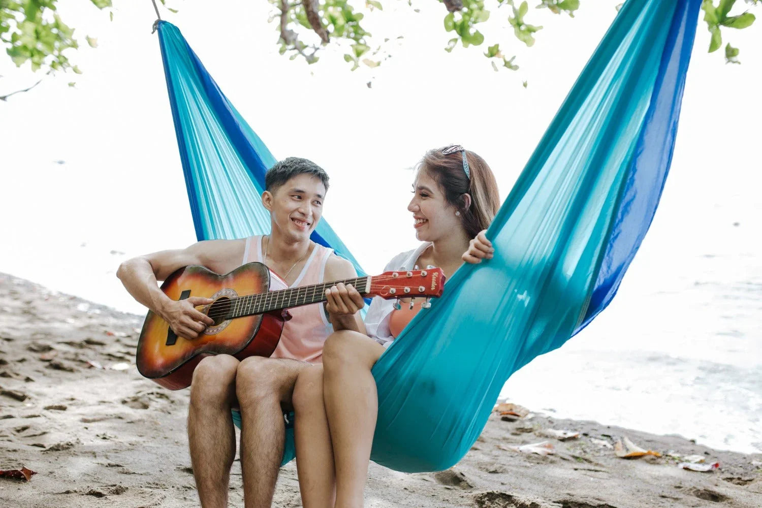 Couple relaxing in blue hammock on beach, man playing acoustic guitar, summer vacation