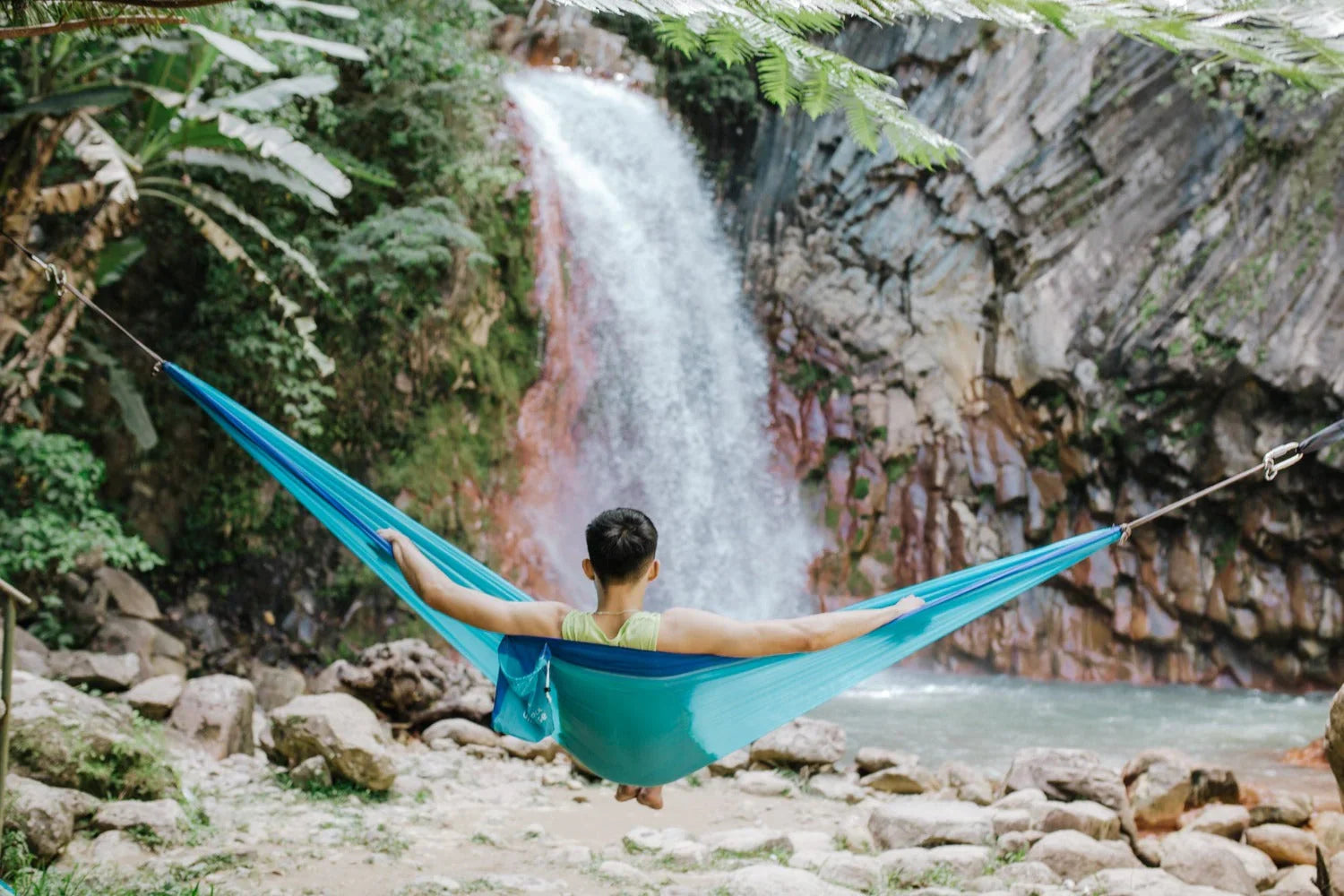 Man relaxing in blue hammock near scenic jungle waterfall and rocky riverbank