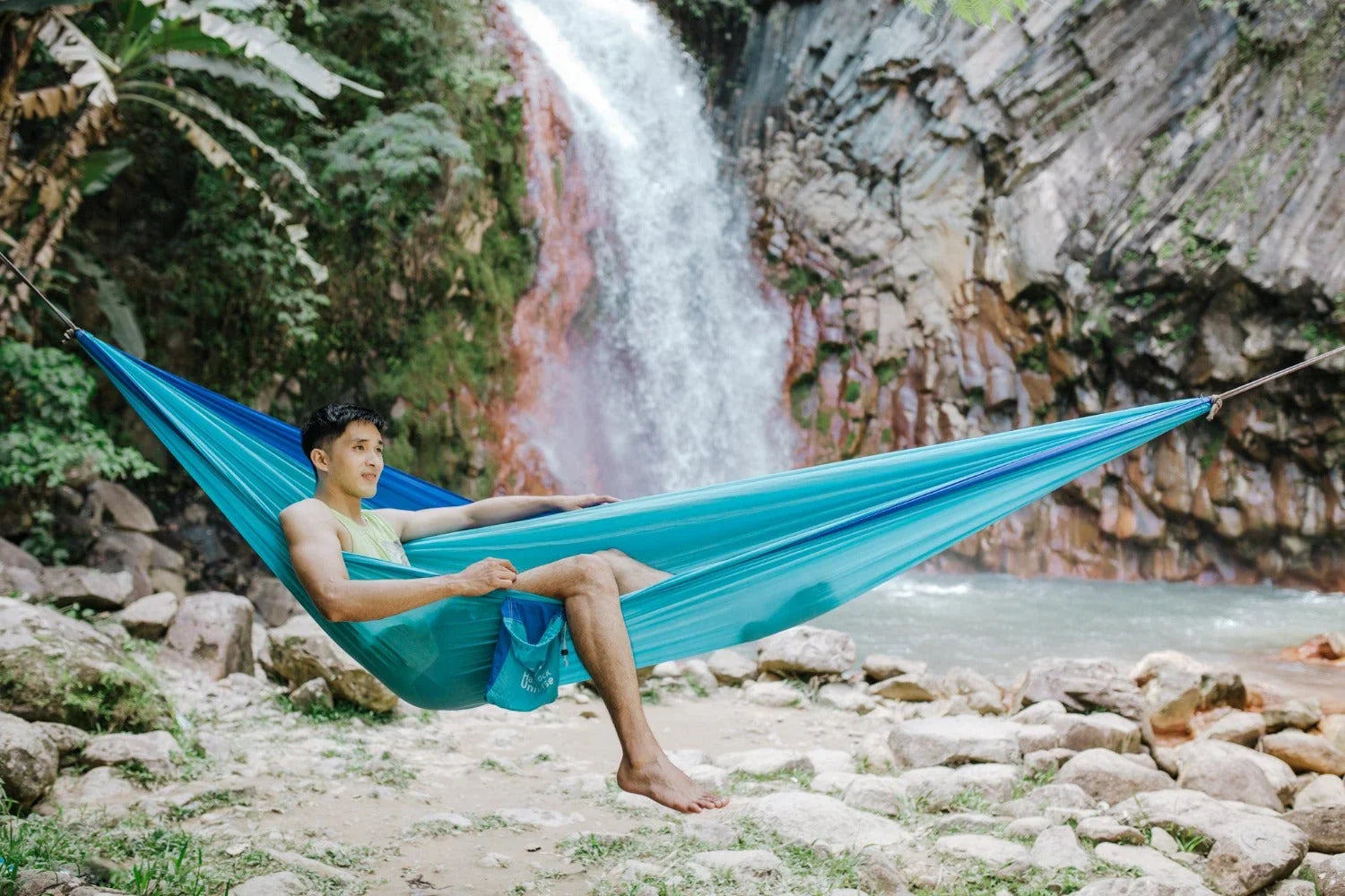 Man relaxing in a blue hammock by a waterfall and rocky riverbank outdoors