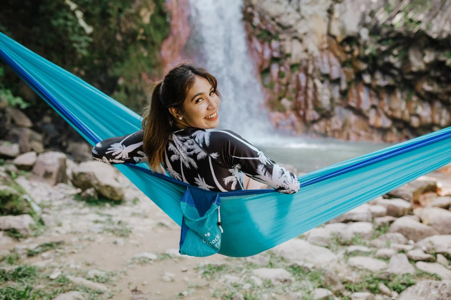 Woman smiling in blue hammock near waterfall outdoors, rocky landscape