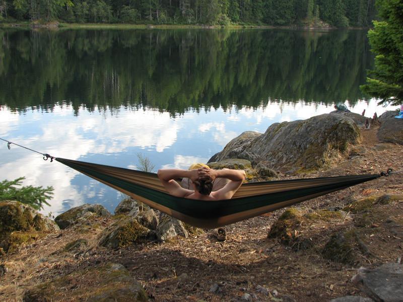Man relaxing in a parachute hammock by a lakeside forest campsite