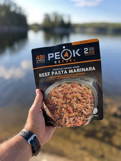 Hand holding Peak Refuel Beef Pasta Marinara meal by a lake in the outdoors
