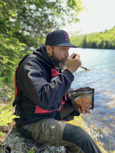 Man in outdoor gear eats beef pasta marinara by a forest lake, enjoying wilderness camping food.