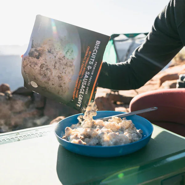 Camping breakfast with biscuits and sausage gravy being served into a blue bowl outdoors