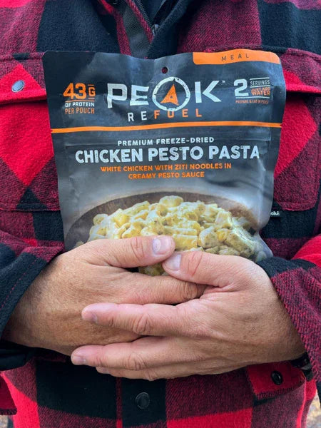 Hands holding Peak Refuel chicken pesto pasta freeze-dried meal in plaid shirt outdoors.