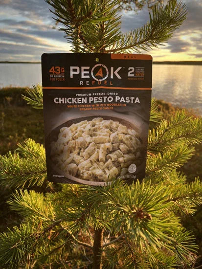Peak Refuel Chicken Pesto Pasta freeze-dried meal on pine tree outdoors by lake at sunset.