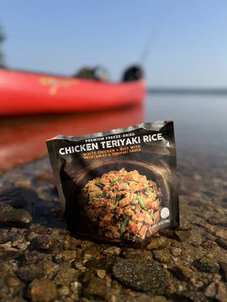 Chicken Teriyaki Rice freeze-dried meal pack on lakeshore with red kayak in background