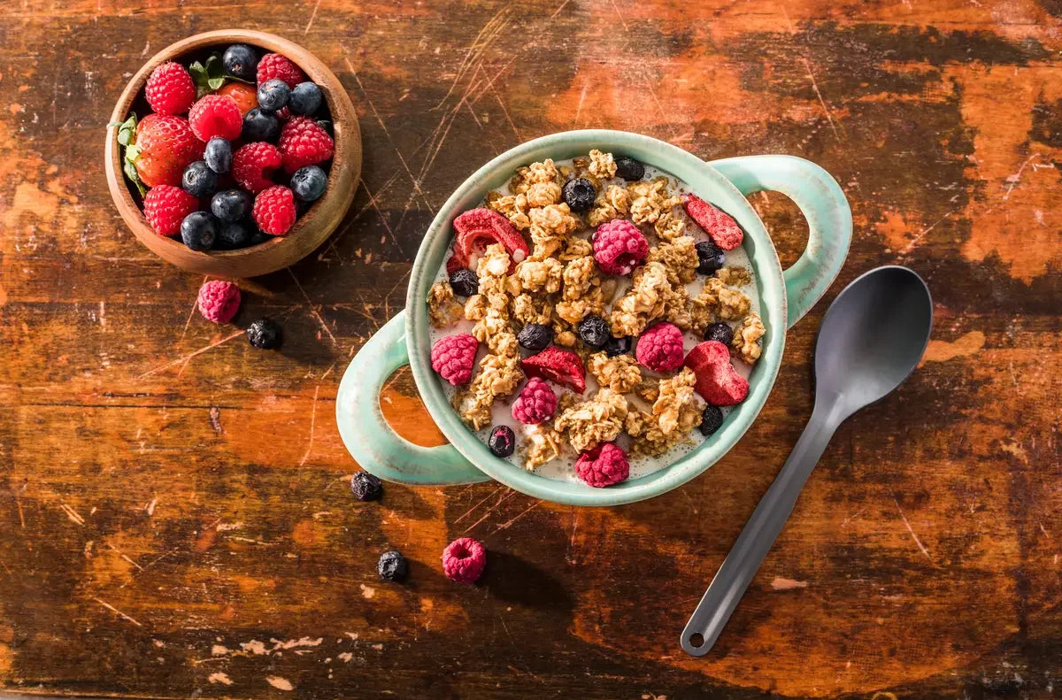 Granola with berries in a green bowl and a wooden bowl of mixed berries on rustic wood table