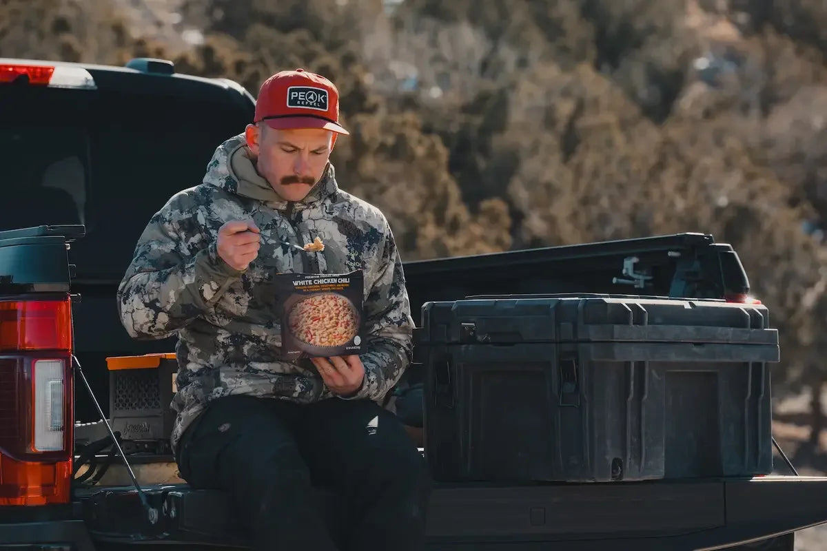 Man in camo jacket eating Peak Refuel white chicken chili on truck tailgate outdoors
