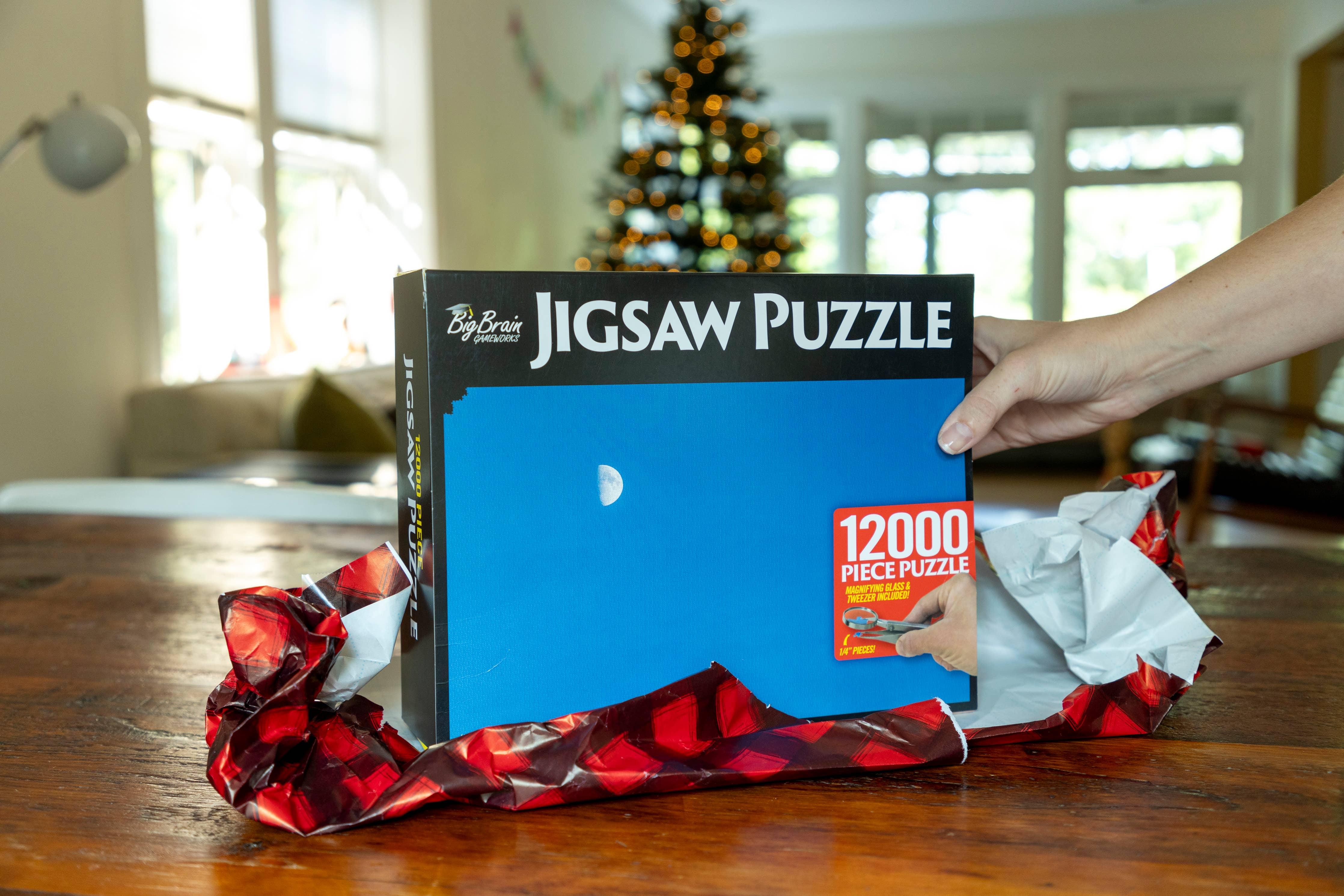 Hand opening 12000 piece jigsaw puzzle box with blue cover on wooden table, Christmas tree in background