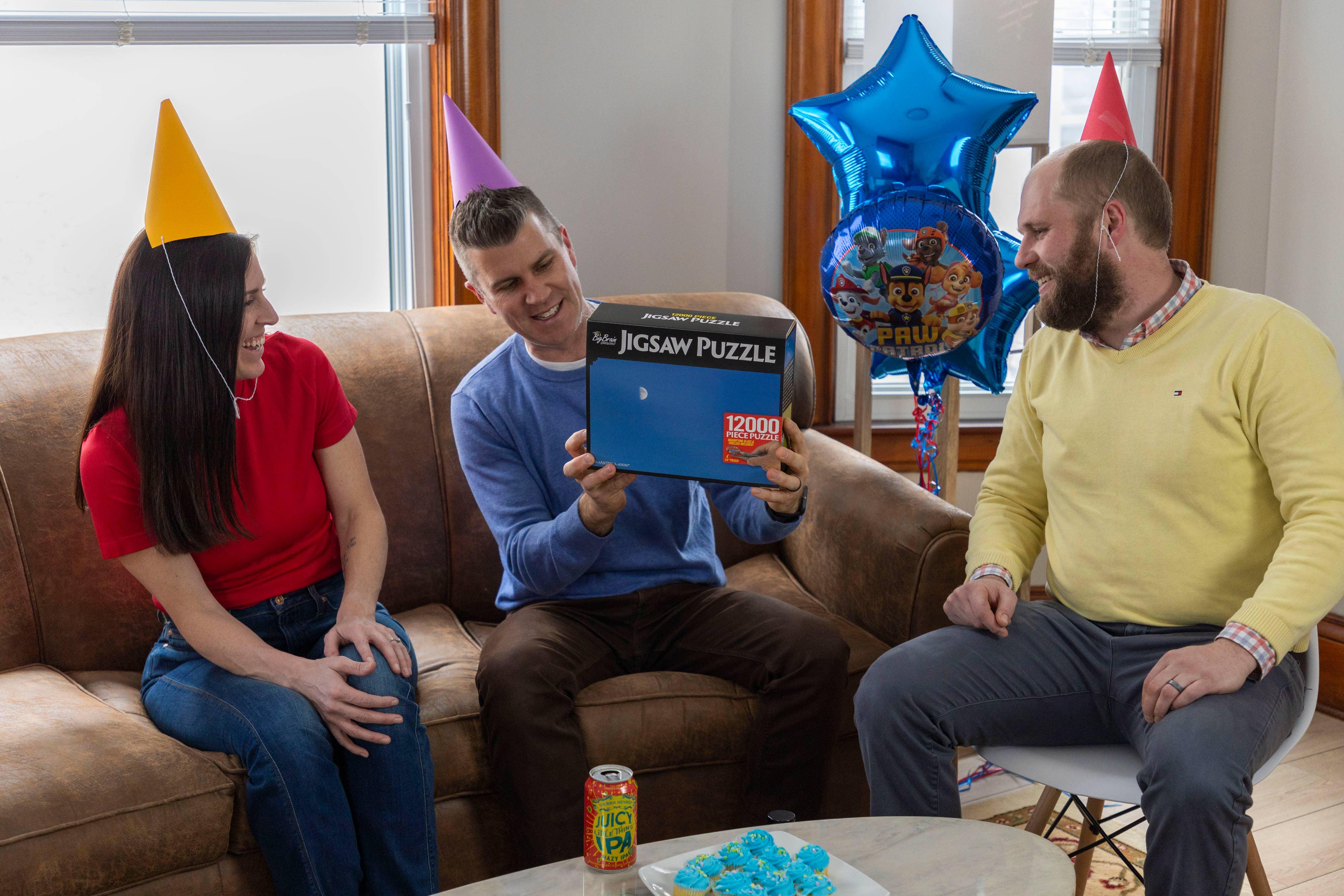 Three adults with party hats celebrate with a 12000 piece jigsaw puzzle gift and blue balloons