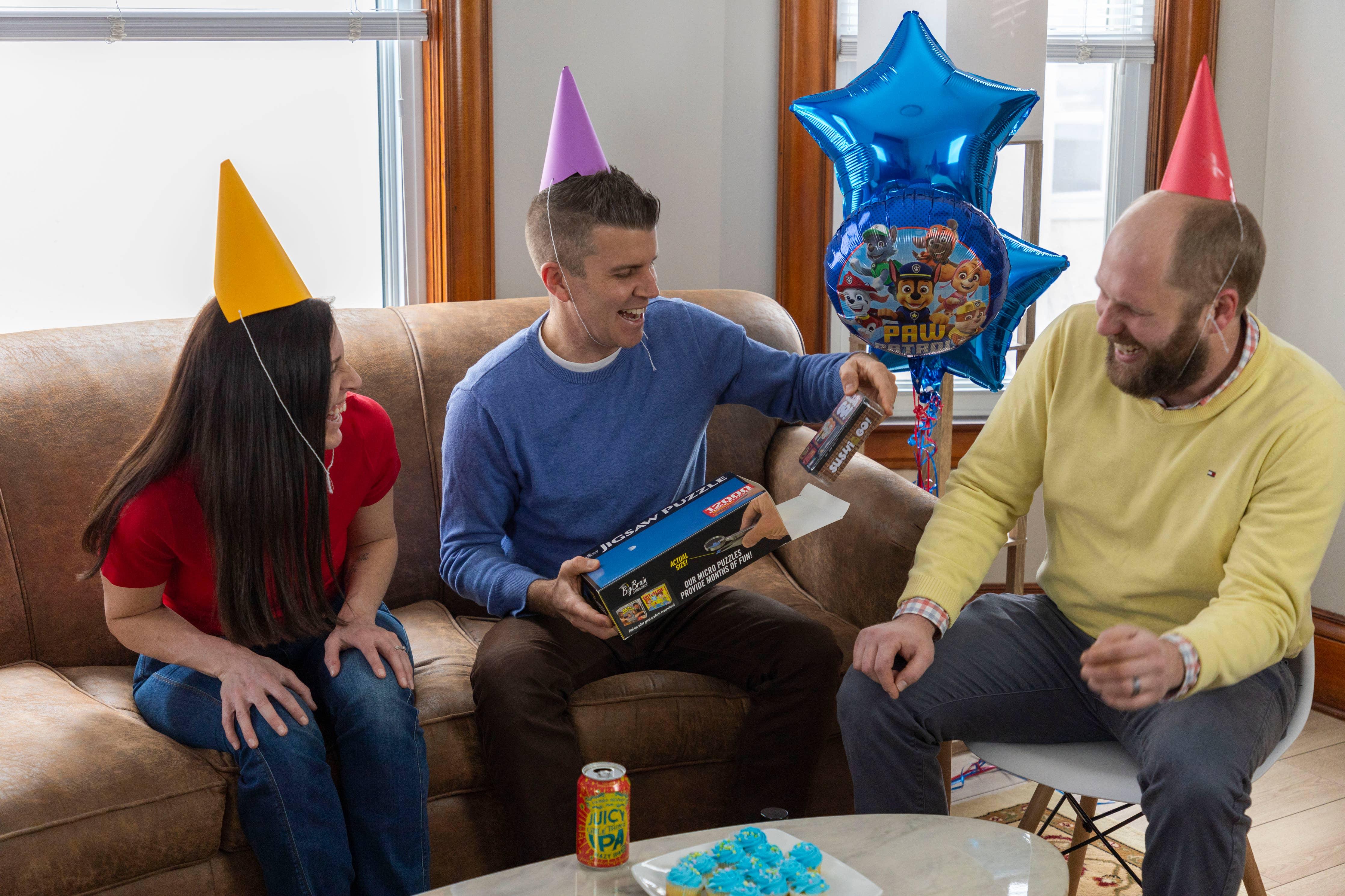 Three adults wearing party hats laugh on a sofa with party decorations and a Paw Patrol balloon.