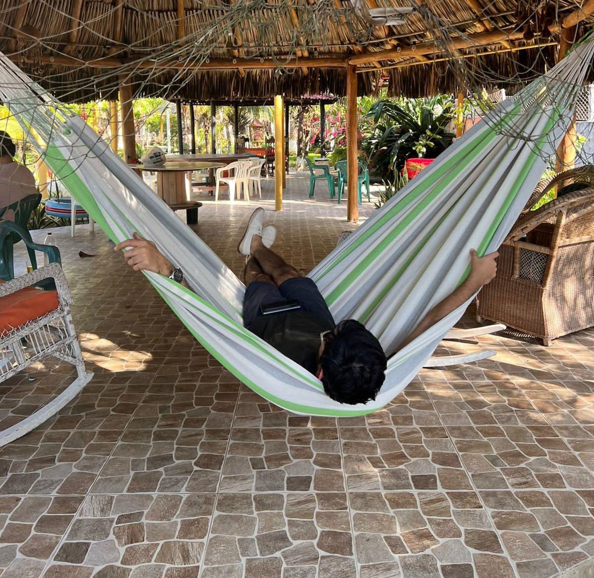 Person relaxing in green, grey, and white Brazilian hammock under thatched patio
