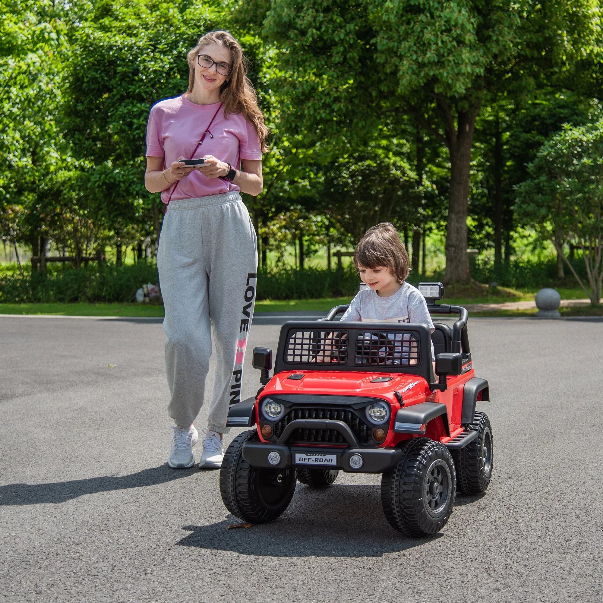 Child driving red ride-on toy car outdoors with adult woman walking beside