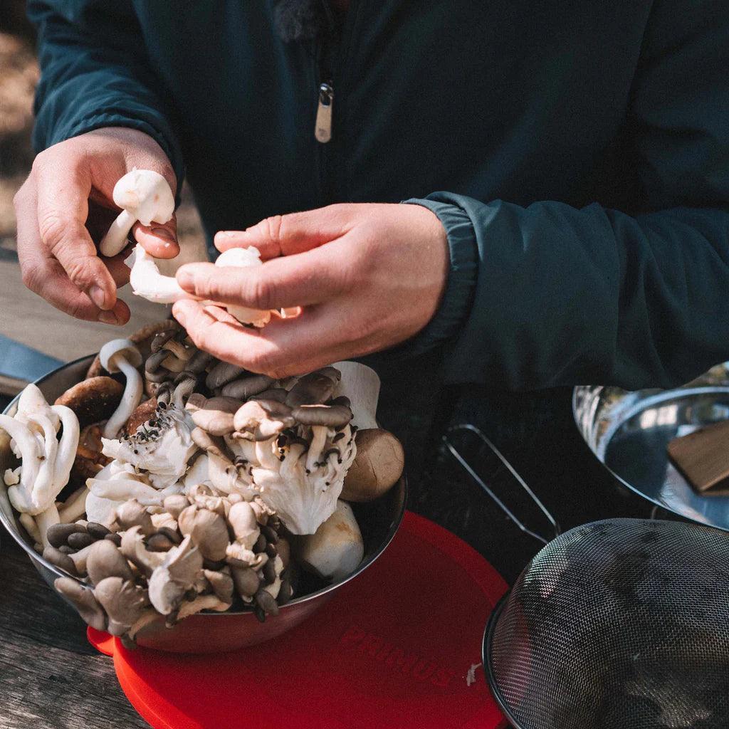 Hands preparing fresh wild mushrooms in a Primus stainless steel campfire bowl outdoors