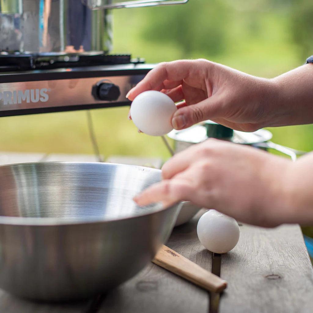 Hands cracking eggs into a stainless steel Primus campfire bowl outdoors on a wooden table