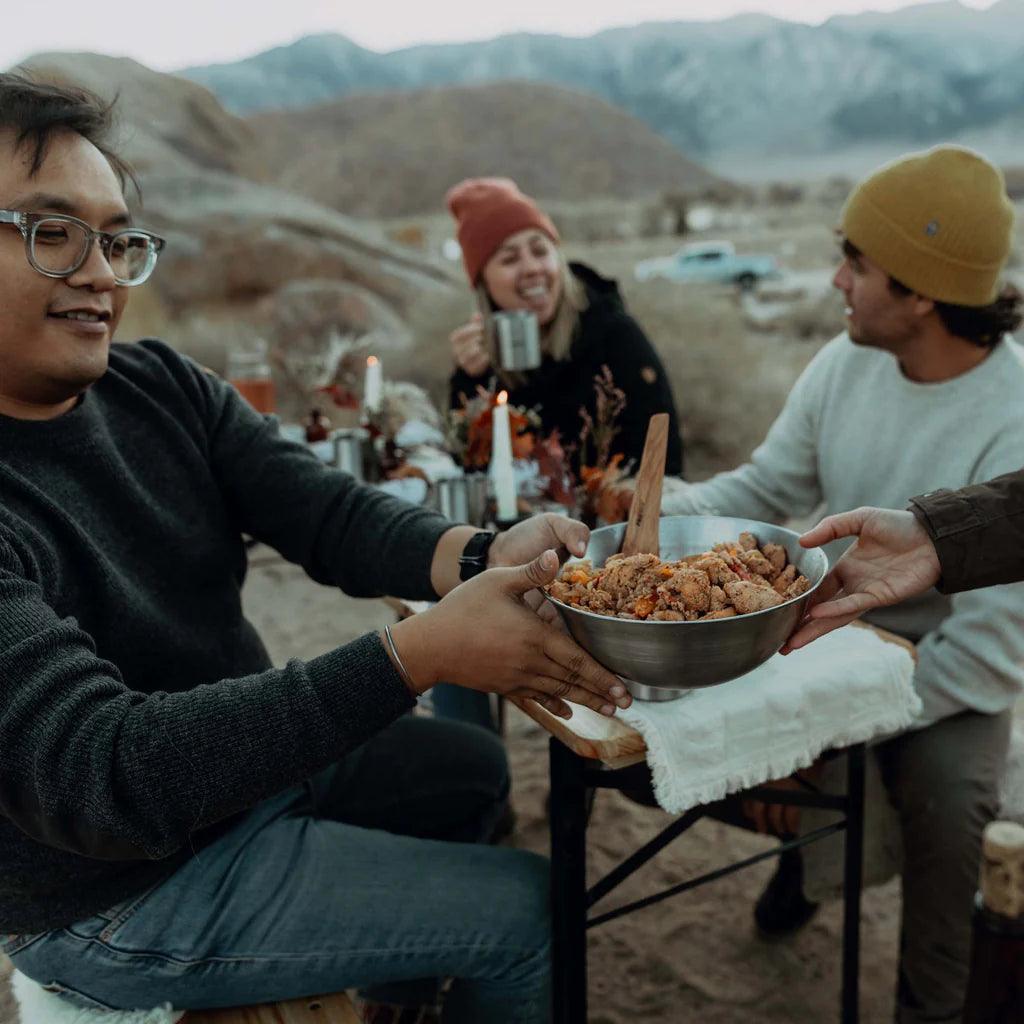 Friends sharing food in a Primus Campfire Bowl at an outdoor table in the mountains.