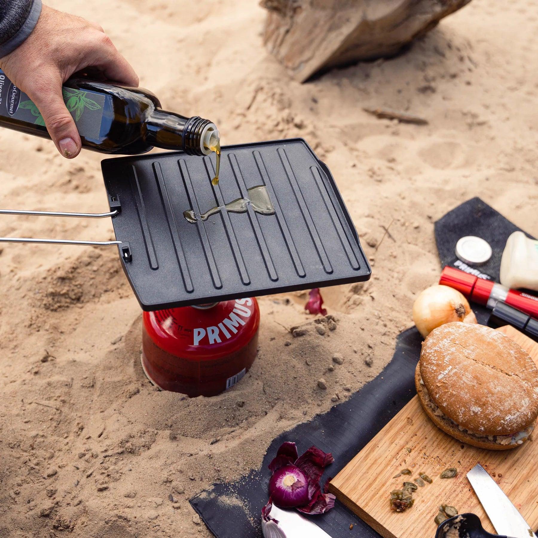 Pouring oil on a Primus campfire griddle plate outdoors with bread and onions nearby