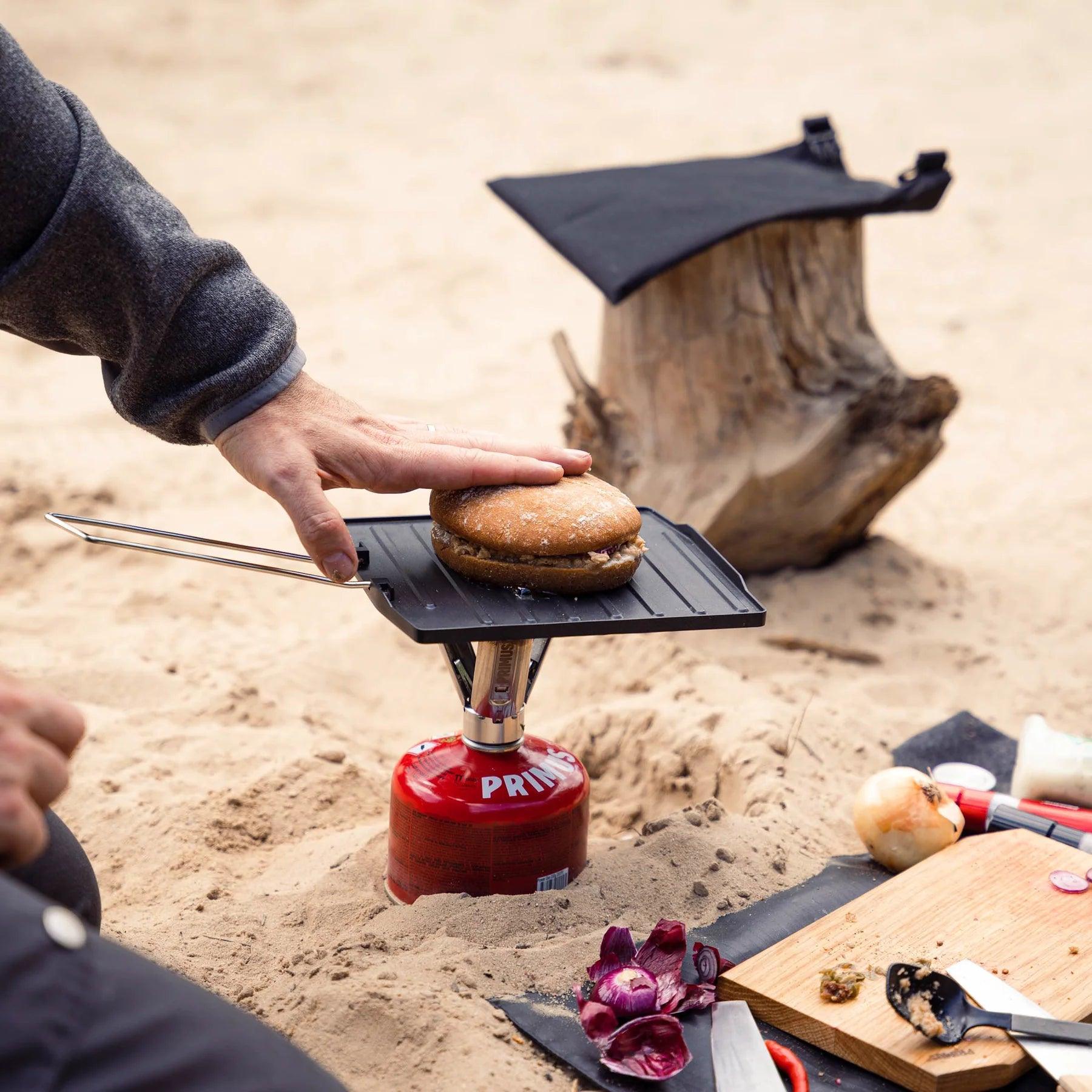 Camping griddle plate on Primus stove cooking burger on sandy outdoor setting