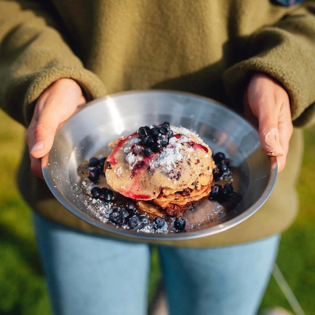 Person holding a metal plate with pancakes topped with blueberries outdoors